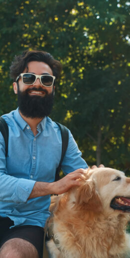 Handsome bearded, stylish man hipster sitting with his dog, golden retriever, on the chair in the park, with trees and sunlight in the background, smiling happy while massaging his joyful pet.